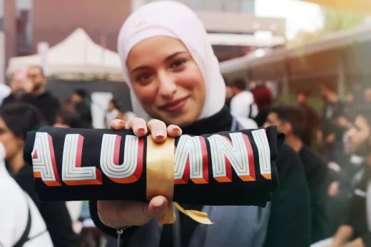 A young woman proudly holding a rolled black fabric tied with a gold ribbon, showcasing the 'ALUMNI' during an AUK event.